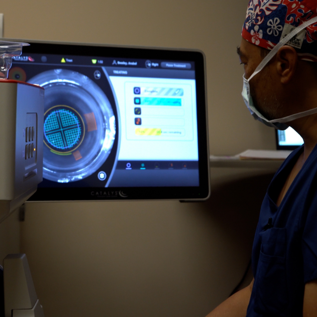 A medical professional wearing scrubs, a surgical mask, and a patterned cap looks at a touchscreen display showing a circular diagnostic or surgical imaging interface in a clinical setting.