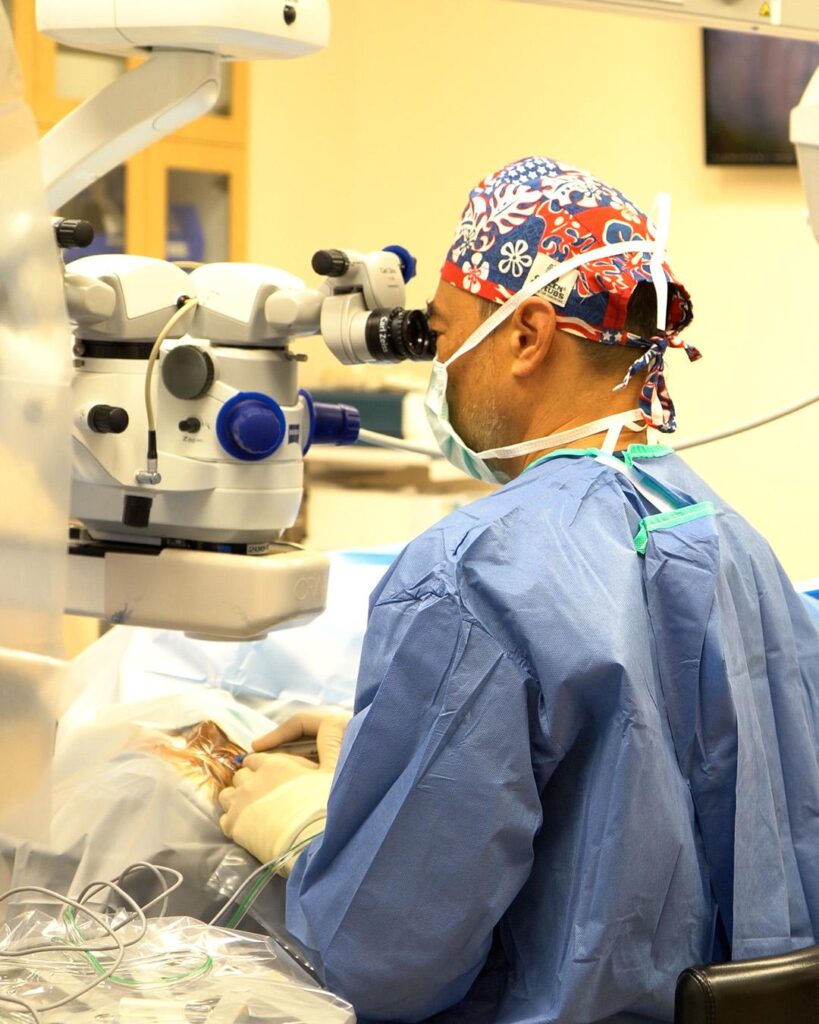 A surgeon wearing a mask, gloves, and a patterned surgical cap operates using a microscope in a brightly lit operating room.